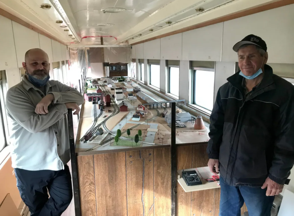 Cory Clark (former executive director) and Doug Peterson (backshop volunteer) stand in front of the displays of replica train models (photo taken by Dog Herbert, CBC)